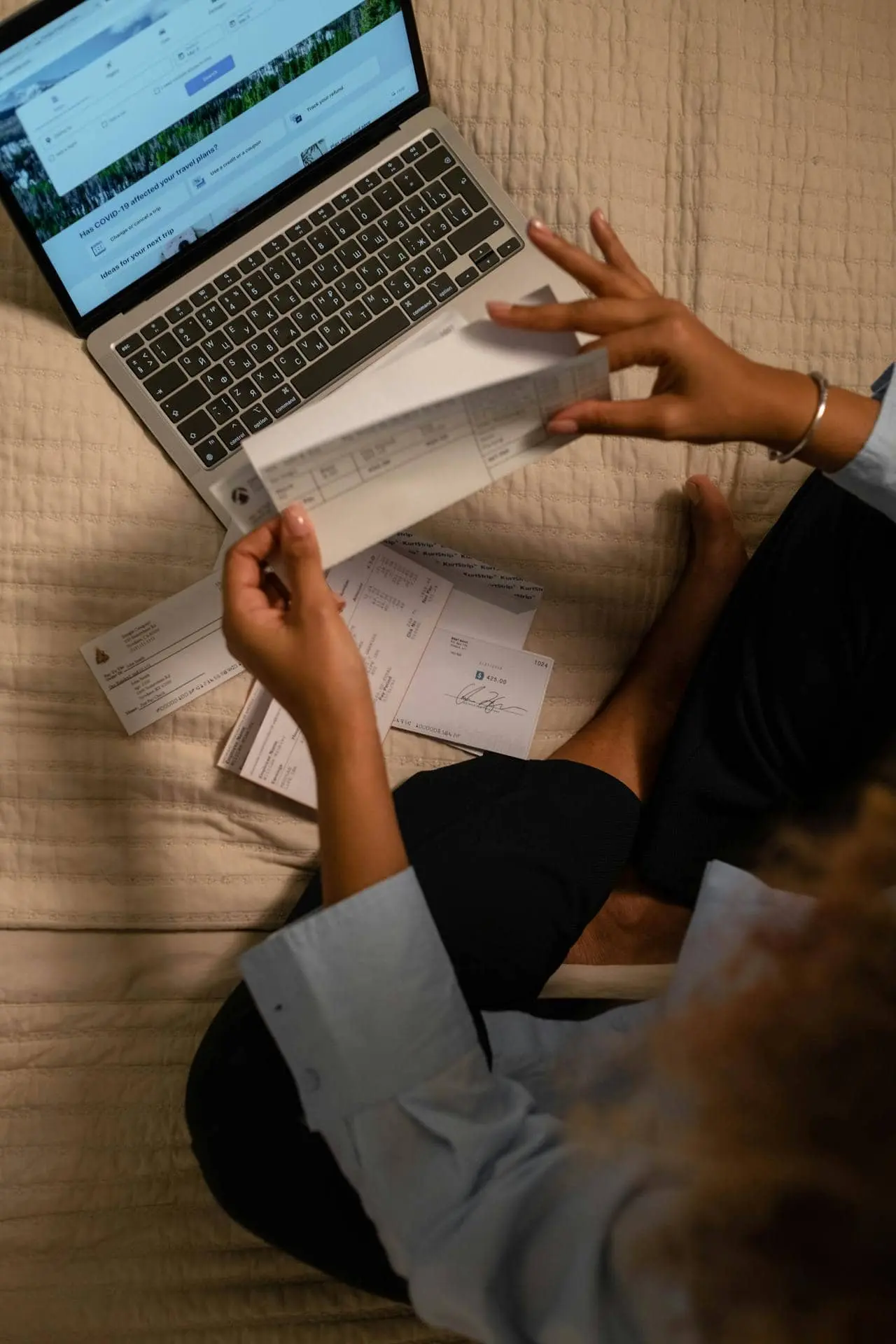 Woman sat on the bed checking paper bills by a laptop